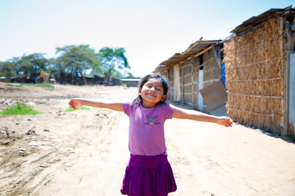 A young Peruvian girl stands in a road with her arms open wide and a smile on her face.