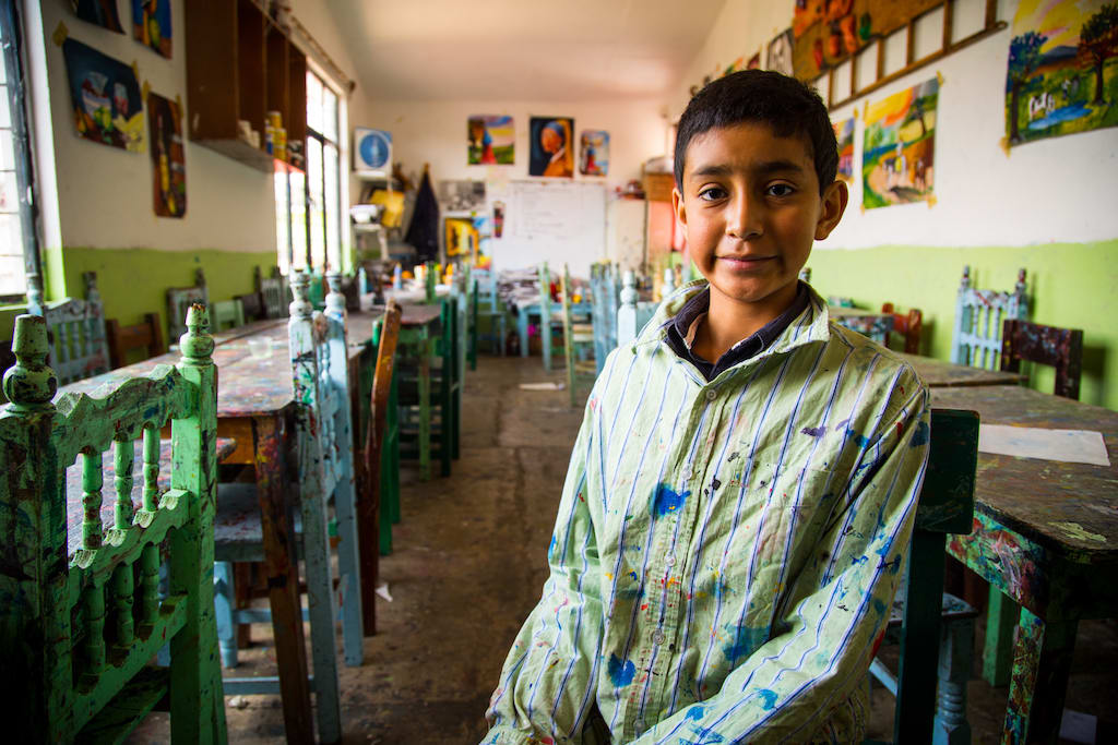 A portrait of Hector sitting in his classroom, wearing his paint smock.