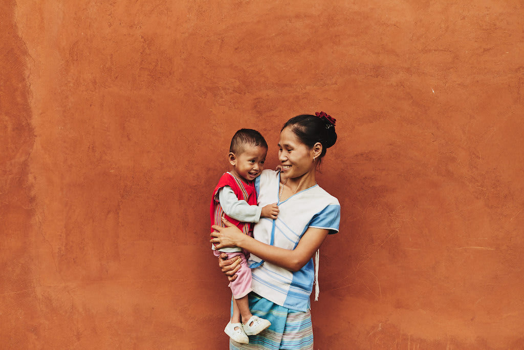 A mother holds a toddler. They are both smiling. They are standing against a rust orange wall.