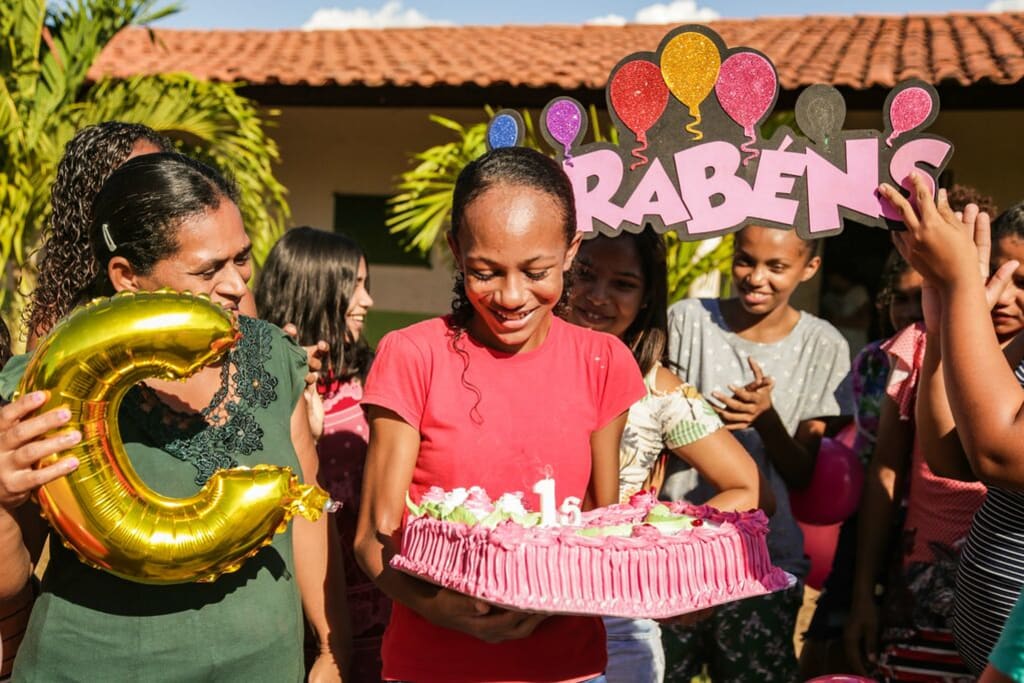 A girl wearing a pink shirt holds a large pink birthday cake. Beside and behind her are balloons.
