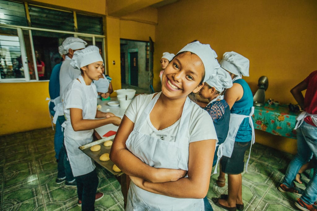A young woman in an apron and chef's hat smiles at the camera as other bakery students work behind her.
