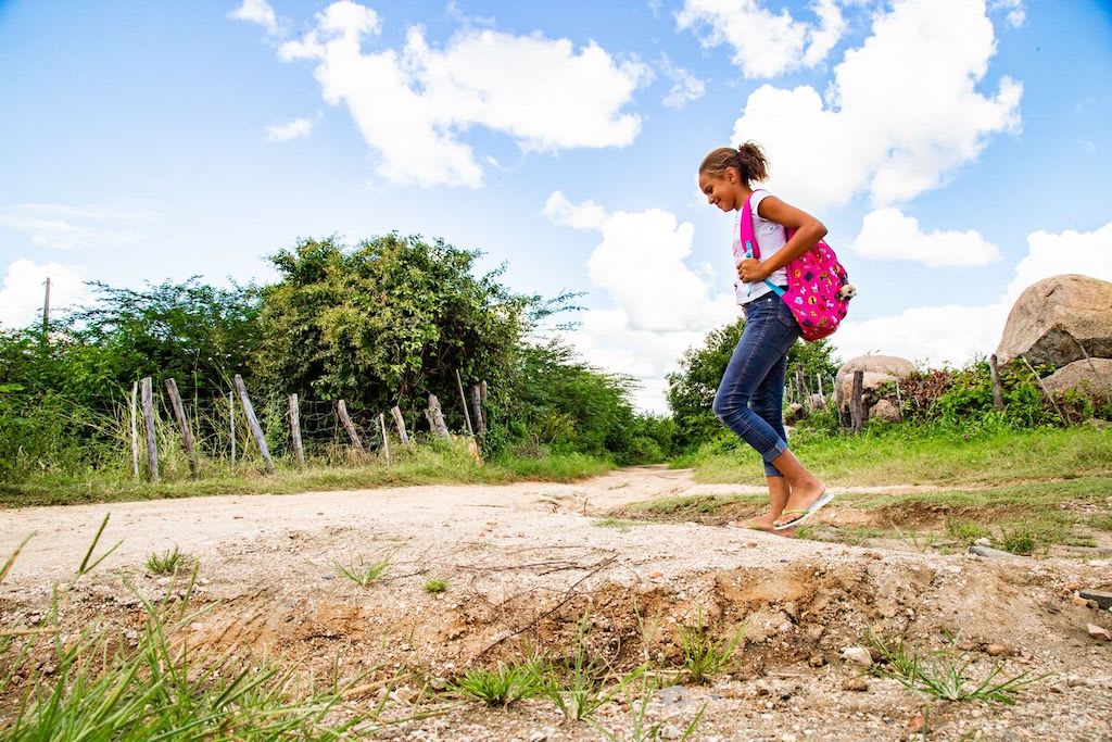 A girl in jeans and a t-shirt, carrying a pink backpack, walks in a field.