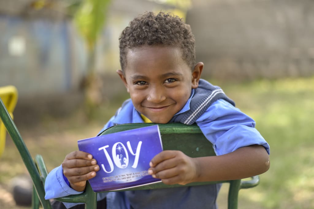 A boy sitting on a chair holds up a Christmas card that says "JOY" on the front.