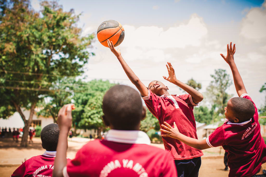 A boy jumps to reach for a basketball, amongst a crowd of other boys engaged in the game.