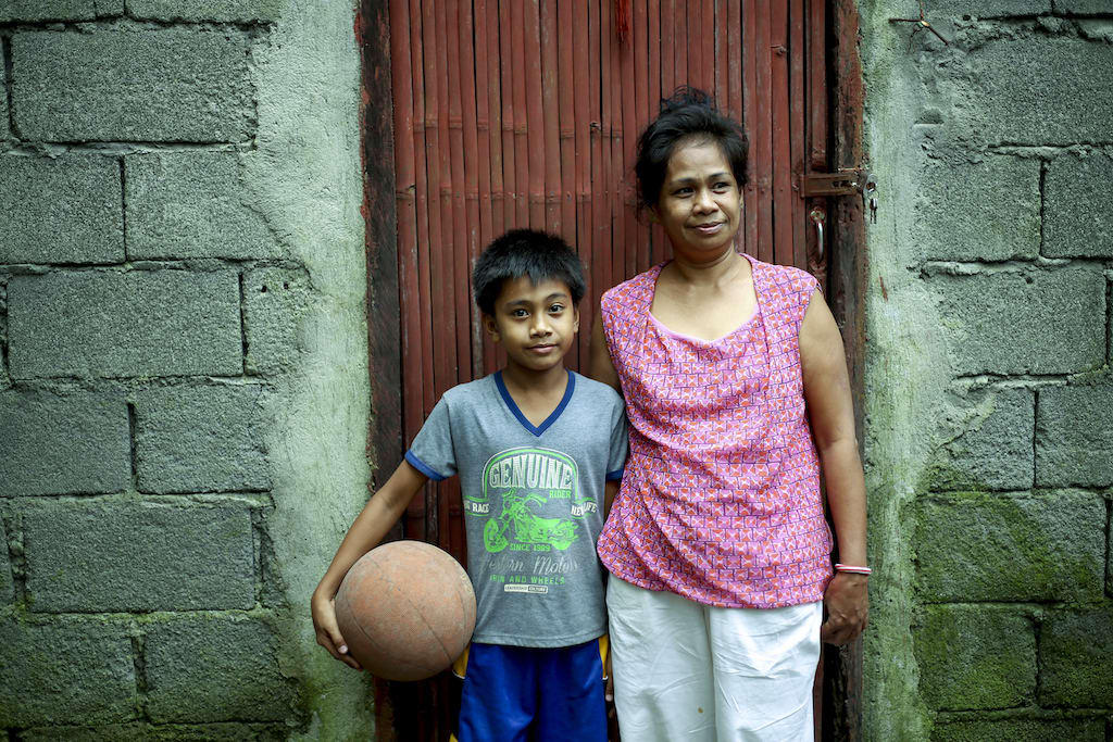 A woman in a pink shirt and a boy in a grey t-shirt stand in front of a brown door to a cinderblock home. The boy is holding a basketball on his hip.