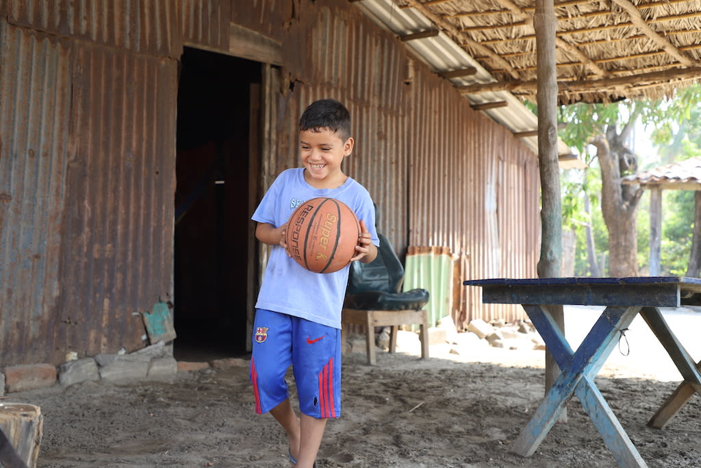A boy in a blue t-shit and blue and red basketball shorts holds a basketball in both hands while walking outside his home.