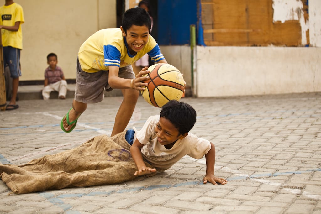 A boy lunges to tag another boy, who is sitting on the ground trying to dodge, with a basketball. They are smiling and laughing.