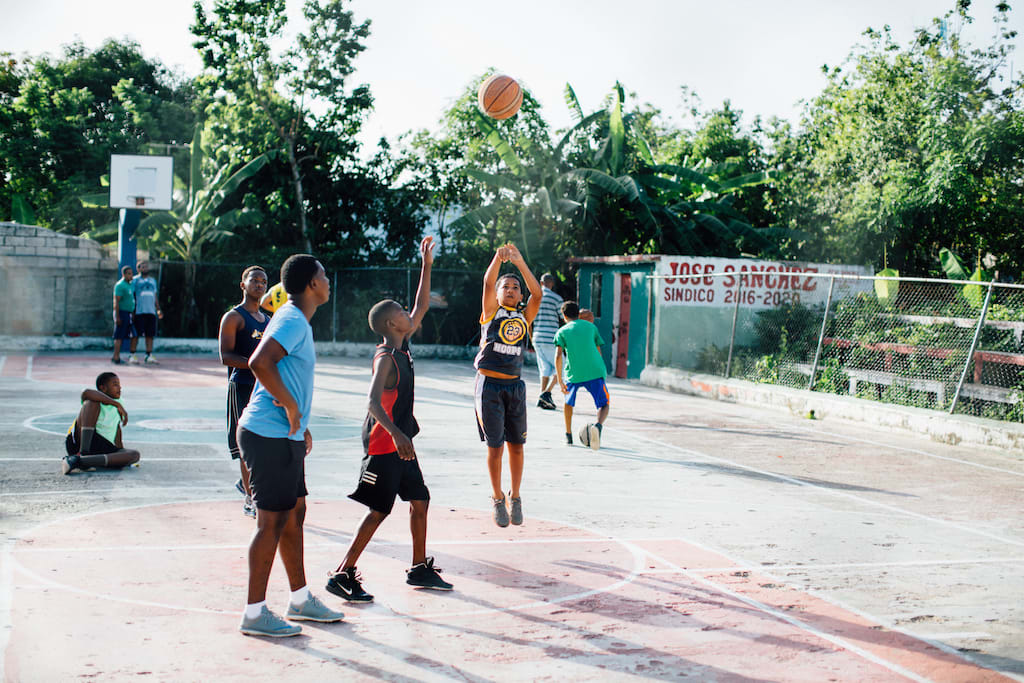 A group of boys play a game of basketball. One boy is shooting the ball while another has his hand up in attempt to block the shot.