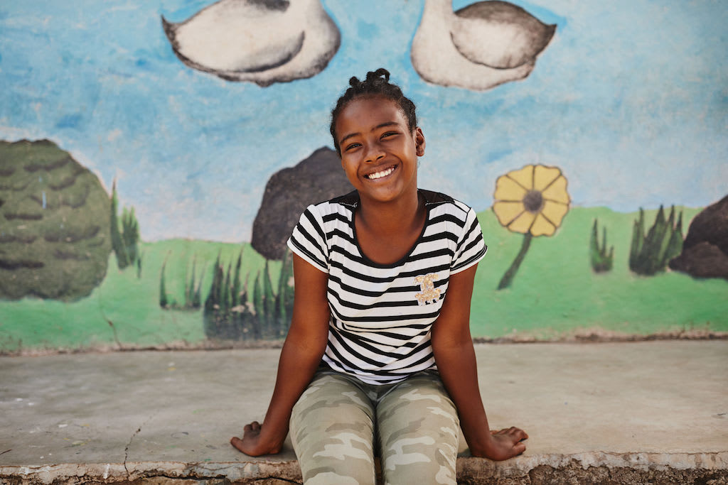 A portrait of Seferash. She is sitting on a step in from of a mural, smiling. She is wearing a black and white stripe tshirt and green camouflage pants.