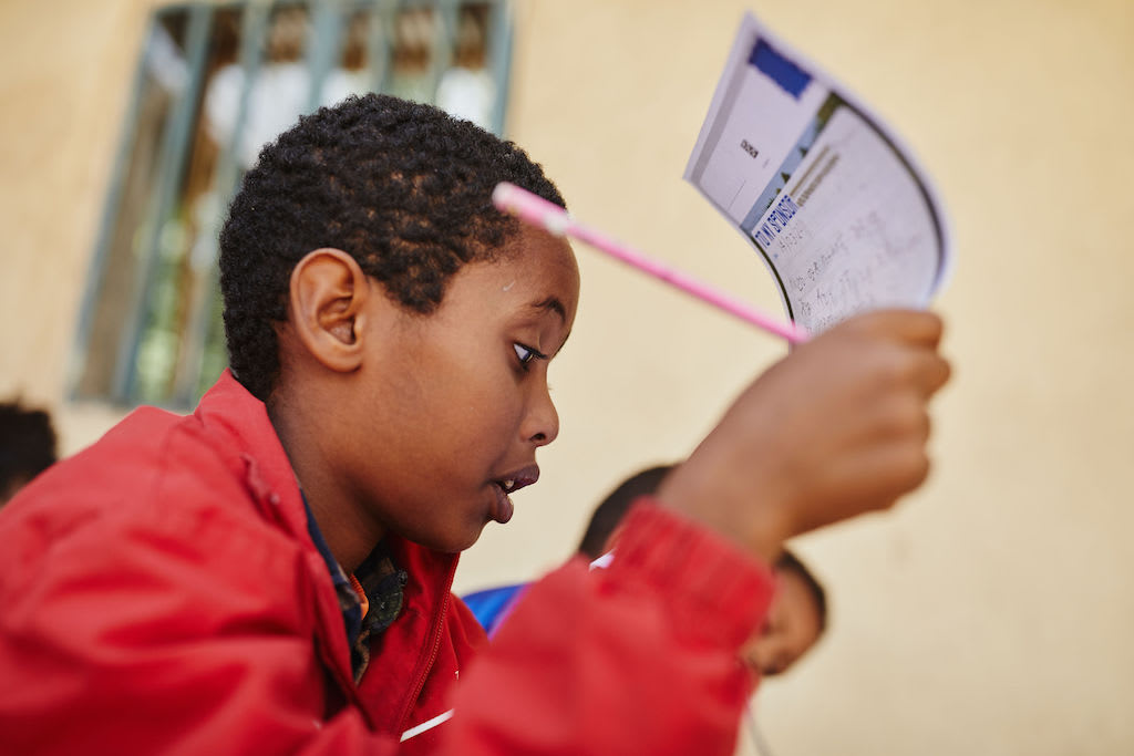While working on a letter to his sponsor, Bemnet holds it up in front of his face to read what he's written. He is wearing a red jacket and holding a pink pencil.