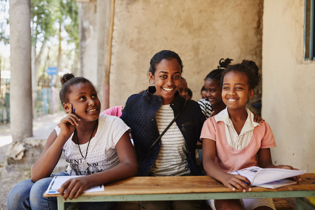 Betey sits at a long desk with two children in the Compassion program as they write letters. They are posing and smiling for the portrait. Betey is wearing a black jacket over a white and black striped t-shirt. The girl on the right is wearing a pink and white top, and the girl on her left is wearing a white t-shirt and blue jeans.