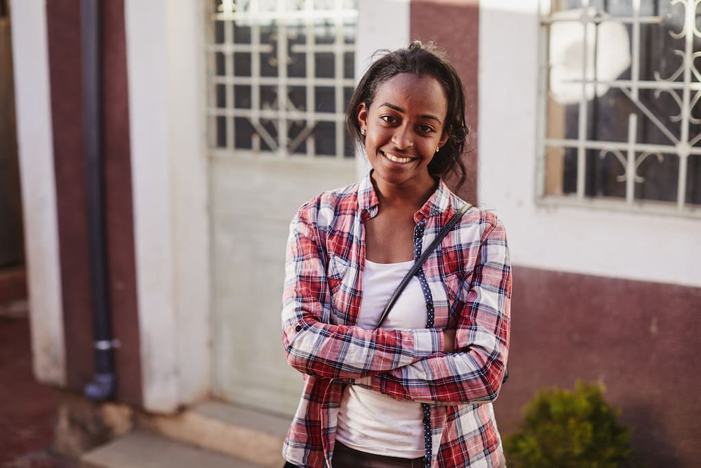 A portrait of Betey. She is wearing a red plaid shirt over a white t-shirt. Her arms are crossed.