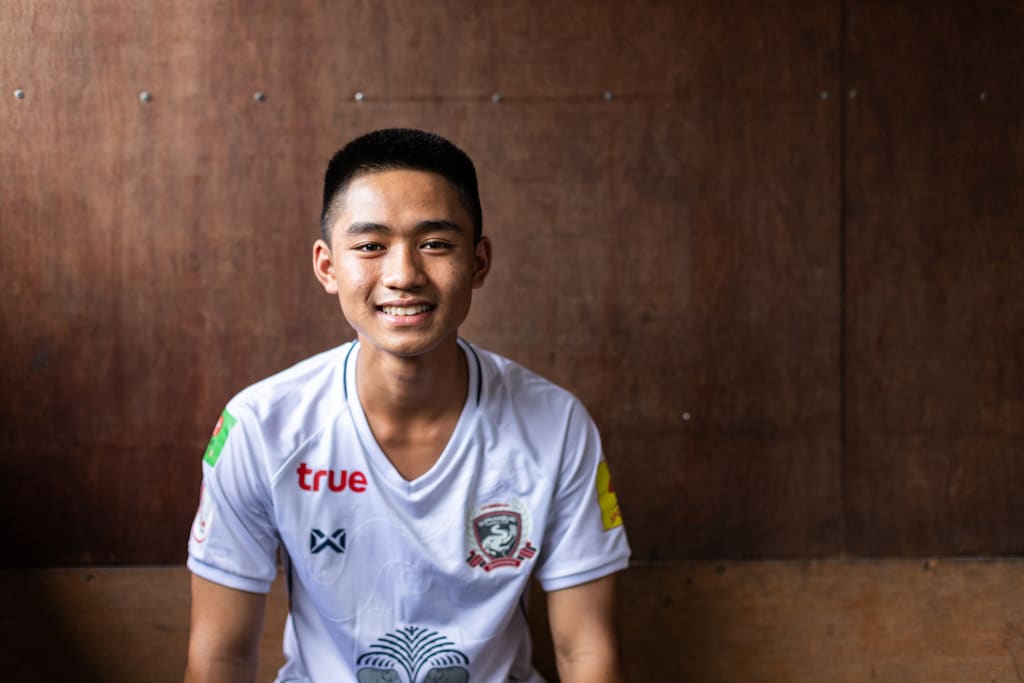 A heard and torso portrait of 15-year-old Adun, member of the soccer team from the Thai cave rescue. He is wearing a white football jersey and looking and smiling straight at the camera.