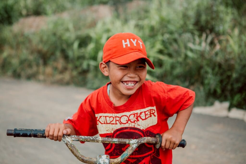 Little boy in an orange shirt and an orange hat grins on his bike