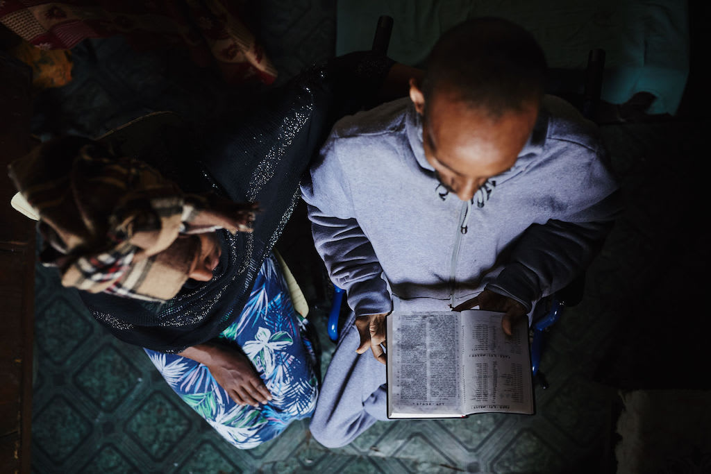 From overhead, a man and woman read the Bible together. The Bible is open on the man's lap as the woman looks over his left shoulder.
