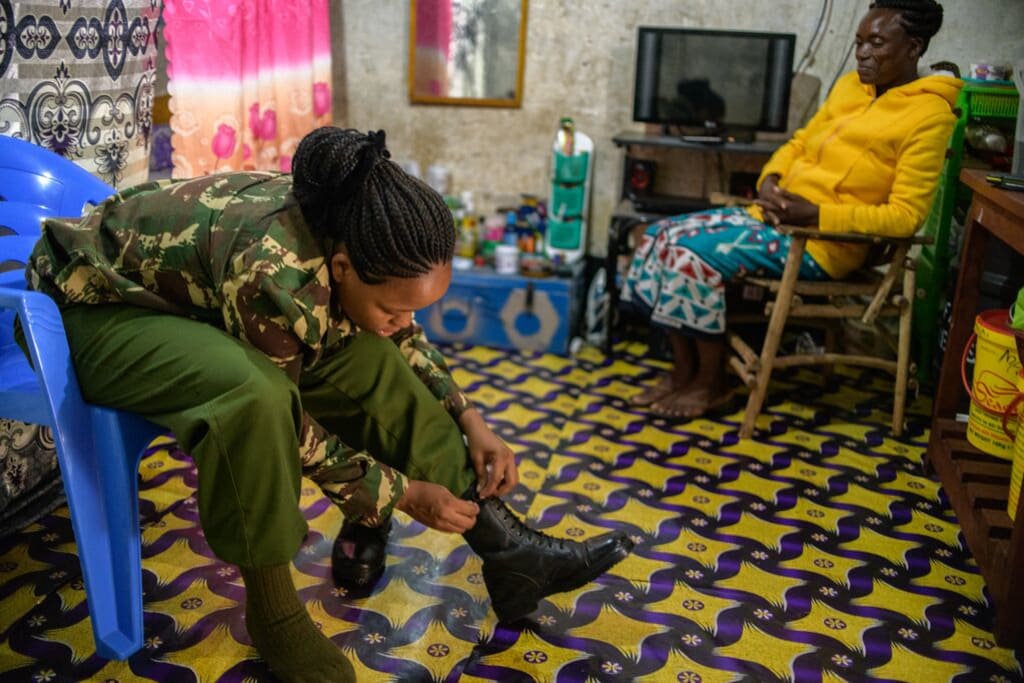 Miriam wearing a camoflauge shirt and green pants. She is sitting in the same room as her mother, who is wearing a yellow sweatshirt. Miriam is lacing up her boots.