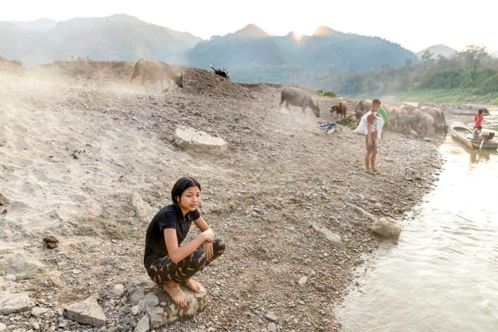 A 15-year-old teenager, female adolescent, Pimchanok is squatting on the beach, shore, sand at the river. Other children, cattle, cows, animals are in the background.