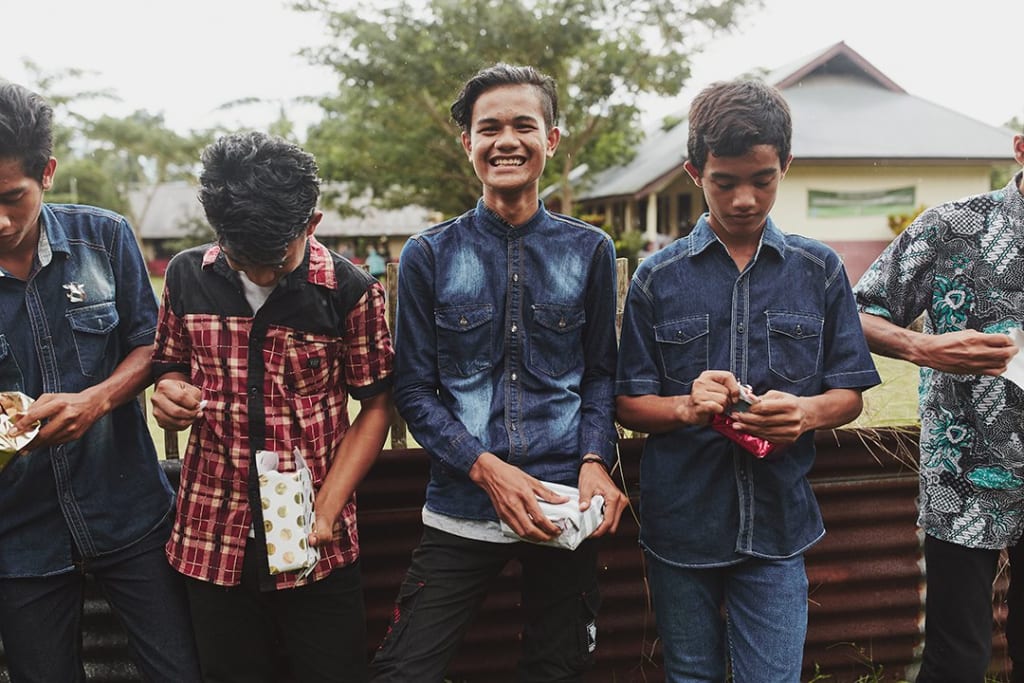 Indonesian, teenage boys stand together opening presents. One smiles as he looks a the camera.