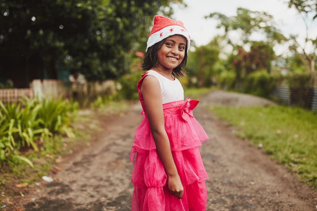 A girl from Indonesia were's a pretty pink dress and a red Christmas hat. She stands in a dirt lane and smiles at the camera.