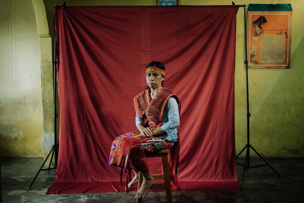 Marhati is sitting in her home in front of a red backdrop. She is wearing traditionally woven clothing.