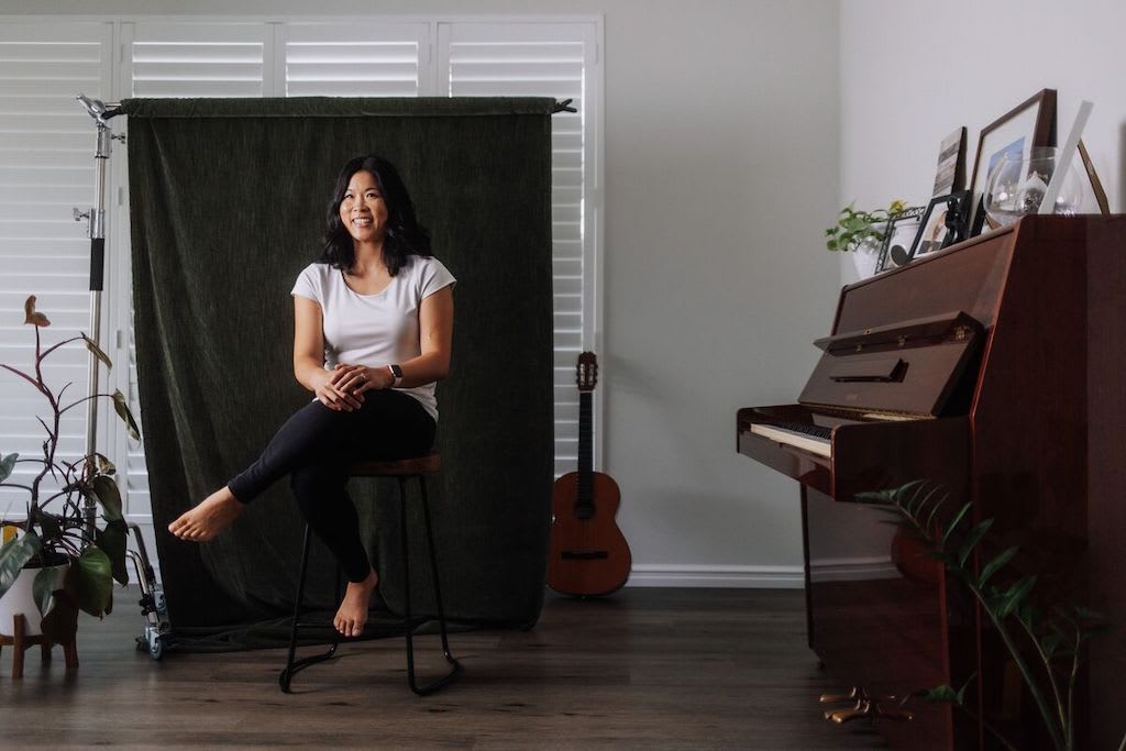 Portrait of Lara in her home. She is wearing a white shirt and black pants. Lara is sitting in front of a green curtain. There is a guitar behind her.