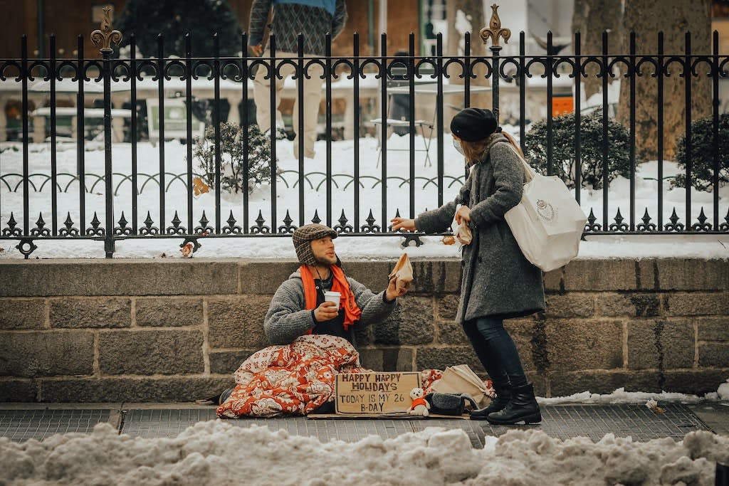A woman hands something to a man sitting on the street.