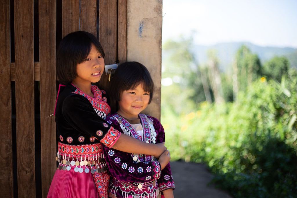 A close up of Nareerart, wearing a maroon traditional Hmong dress. She has her arms around Torfan who’s wearing a purple traditional Hmong dress. They are both standing at the Compassion centre, looking off into the distance and smiling.