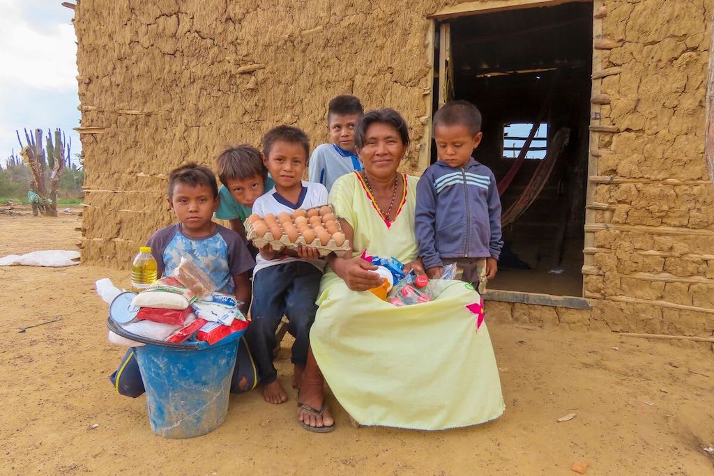 Alirio is wearing a white shirt and holding a tray of eggs. He is sitting outside with his mother and his siblings. There is a bucket of food in front of them which was provided by Compassion.