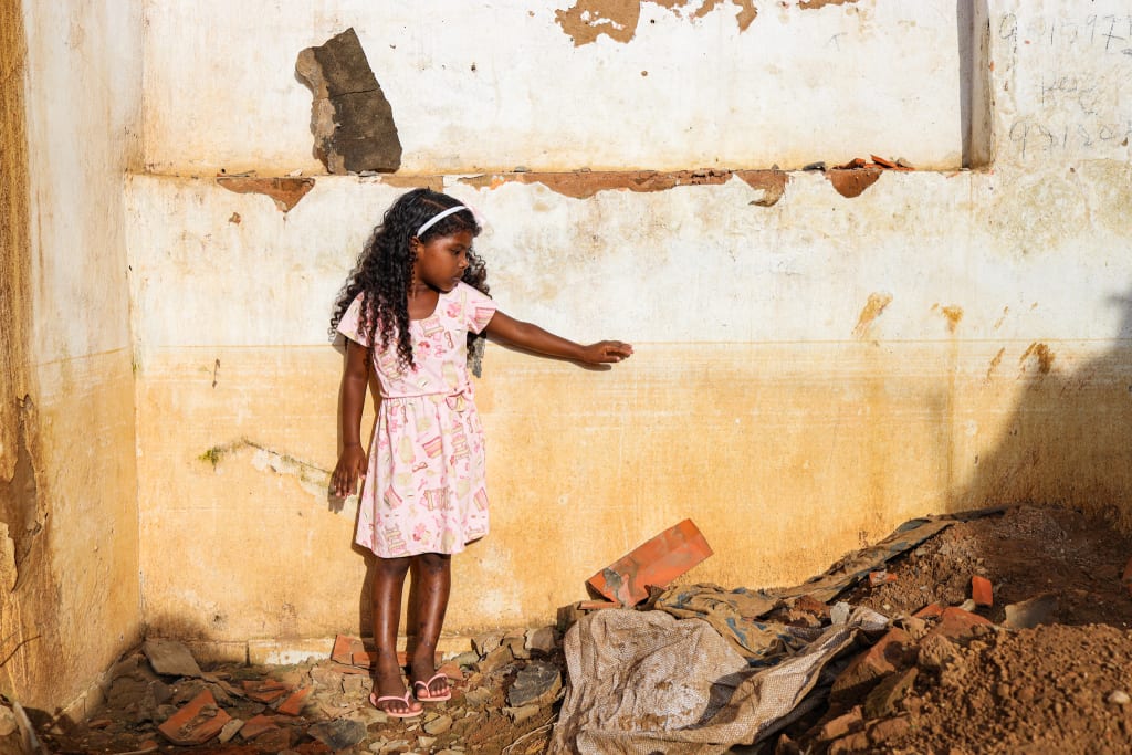 A girl in a pink dress with long black hair stands in front of a stained wall.