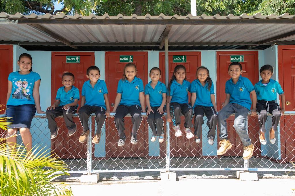 Kids in El Salvador sit on a bench in front of a newly-constructed washroom block in their community.