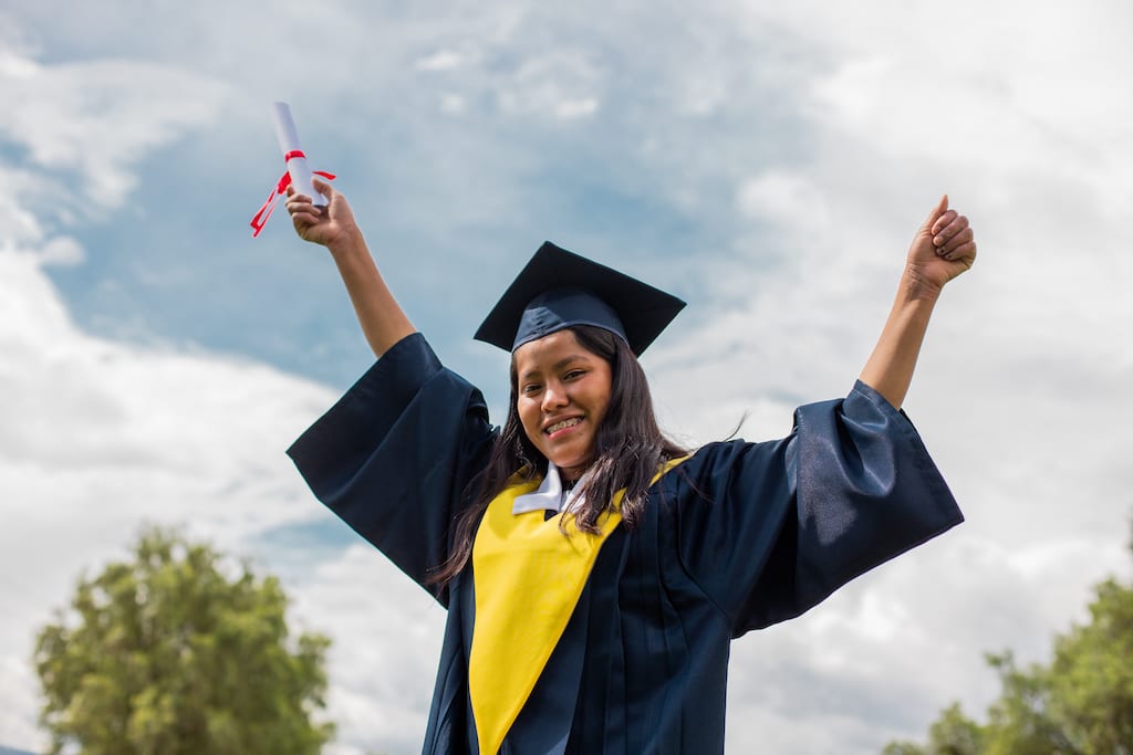 Maribel is wearing her graduation uniform, a black cap and gown, and a yellow collar. She is standing outside with her arms up in the air. In one hand is her diploma.