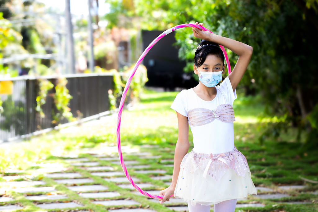 Suleyma is wearing her spinning uniform, a pink and white outfit, and a face mask. She is standing in front of a Compassion office where she is gettign ready to perform. She is holidng a hulu hoop over her head.