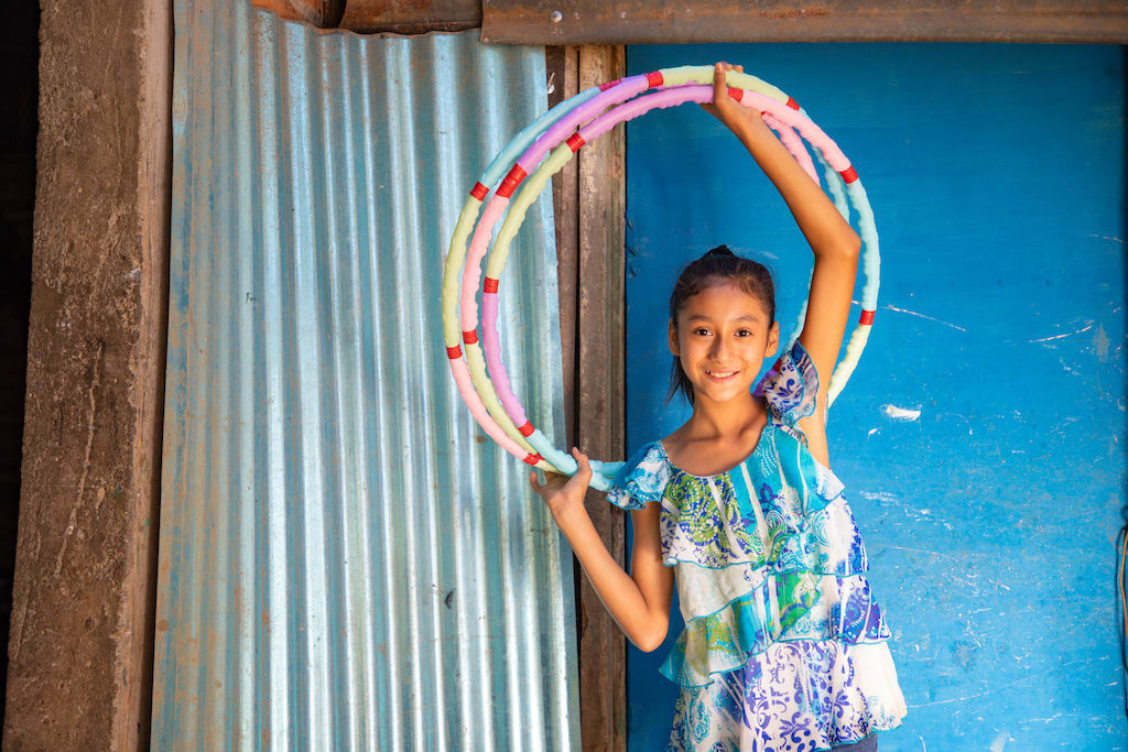 Suleyma is wearing jeans with a colorfully patterned shirt. She is standing in front of her home and is holding three hula hoops above her head. Her home is blue and the door is made of corrugated metal.