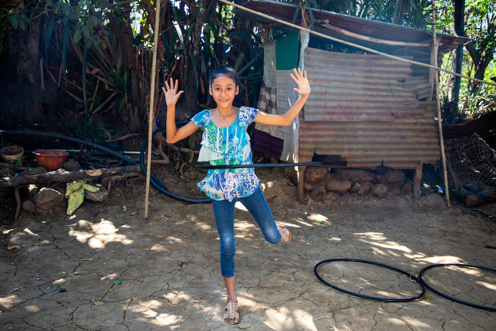Suleyma is wearing jeans with a colorfully patterned shirt. She is standing outside her home and is playing with her hula hoops.