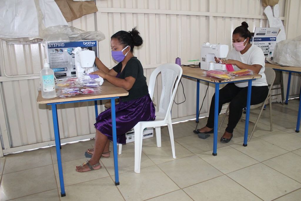 Eveling Sánchez (left) is wearing a black shirt and a purple skirt. Leticia Mayorga (right) is wearing a white shirt and black pants. They are sitting at sewing machines and are making masks.
