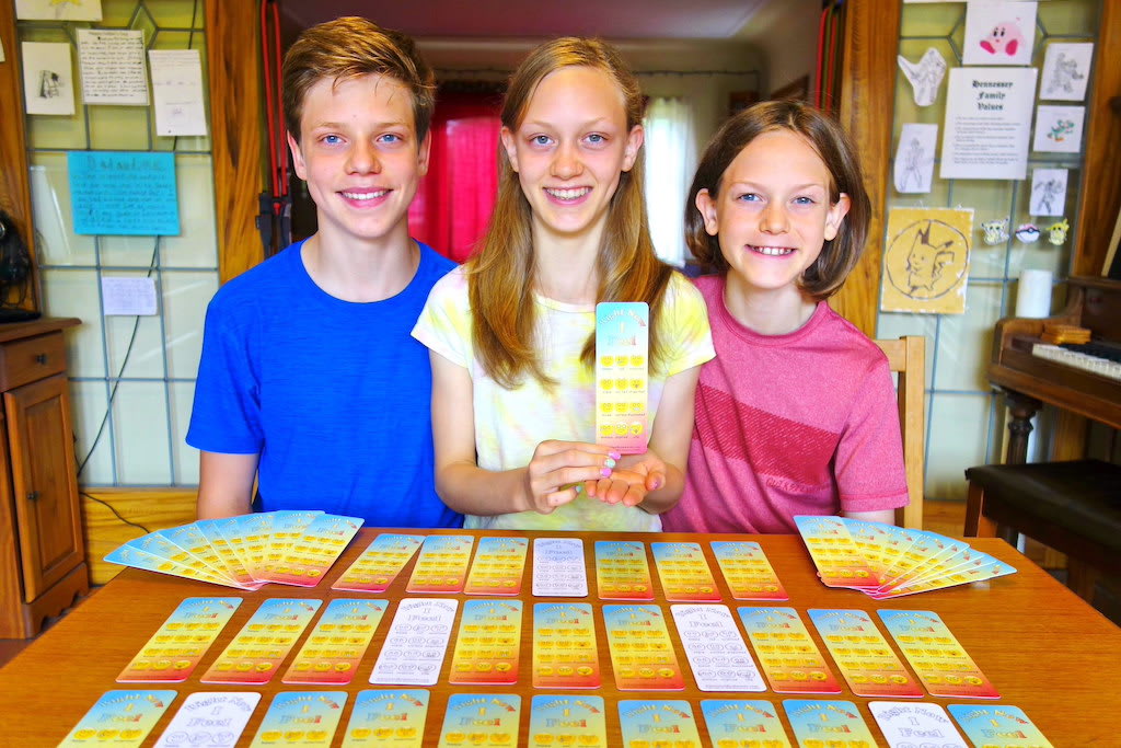 Three siblings sit together at a table with their bookmark product infront of them on the table.