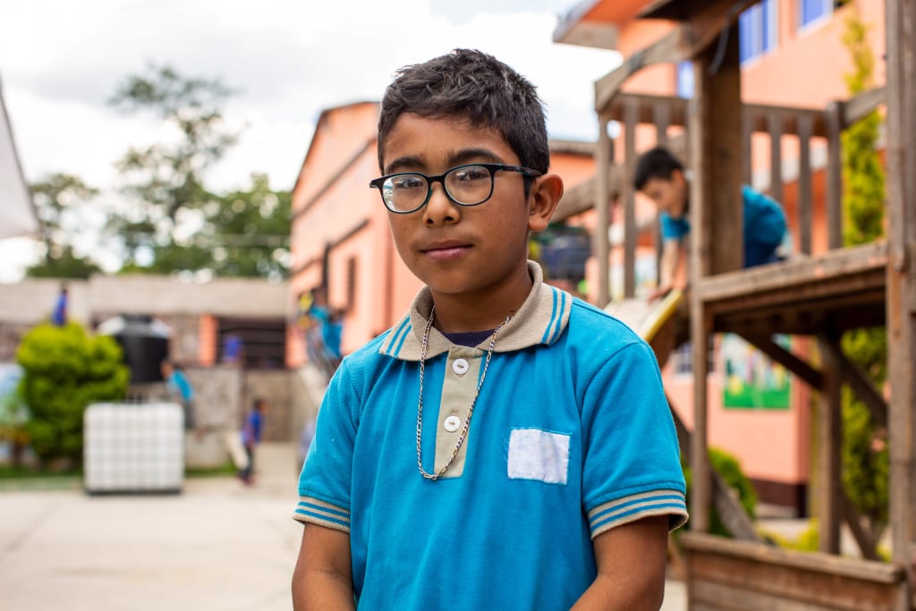 A boy in a blue shirt and glasses looks at the camera. He stands in front of orange buildings.