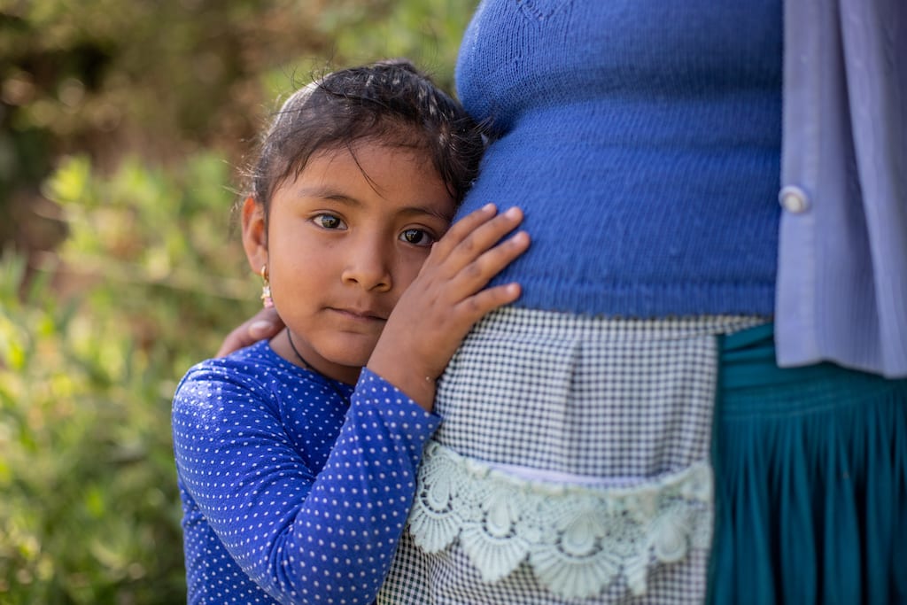 Girl wearing a blue shirt leans against her mom's pregnant stomach.