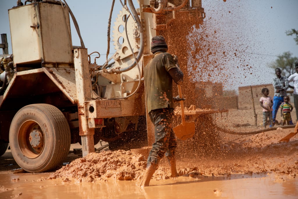 A machine drills a hole in red earth. A man stands with a shovel in front of the machine.