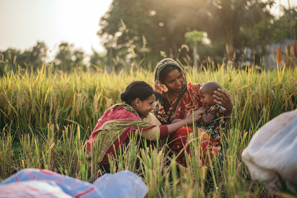 A mother holds her baby in a field in the setting sun. Another girl is leaning over to say hello to the baby. They are all smiling.