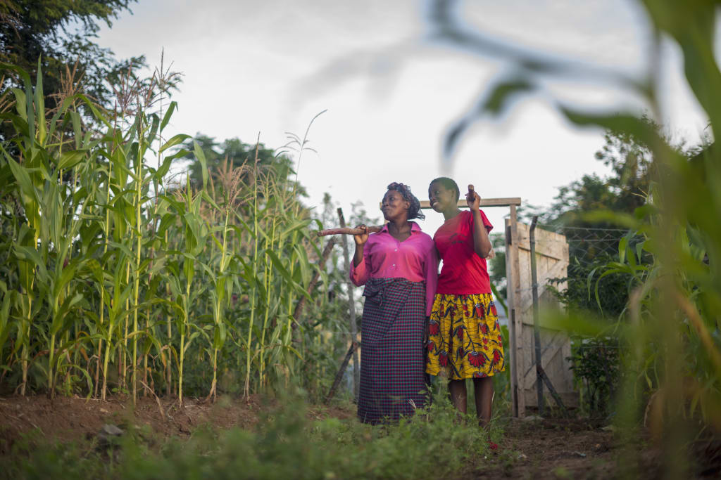 two women stand confidently in a field of corn with gardening tools slung over their shoulders. Miriam and Lilian live in Kenya and have received help from Compassion funds