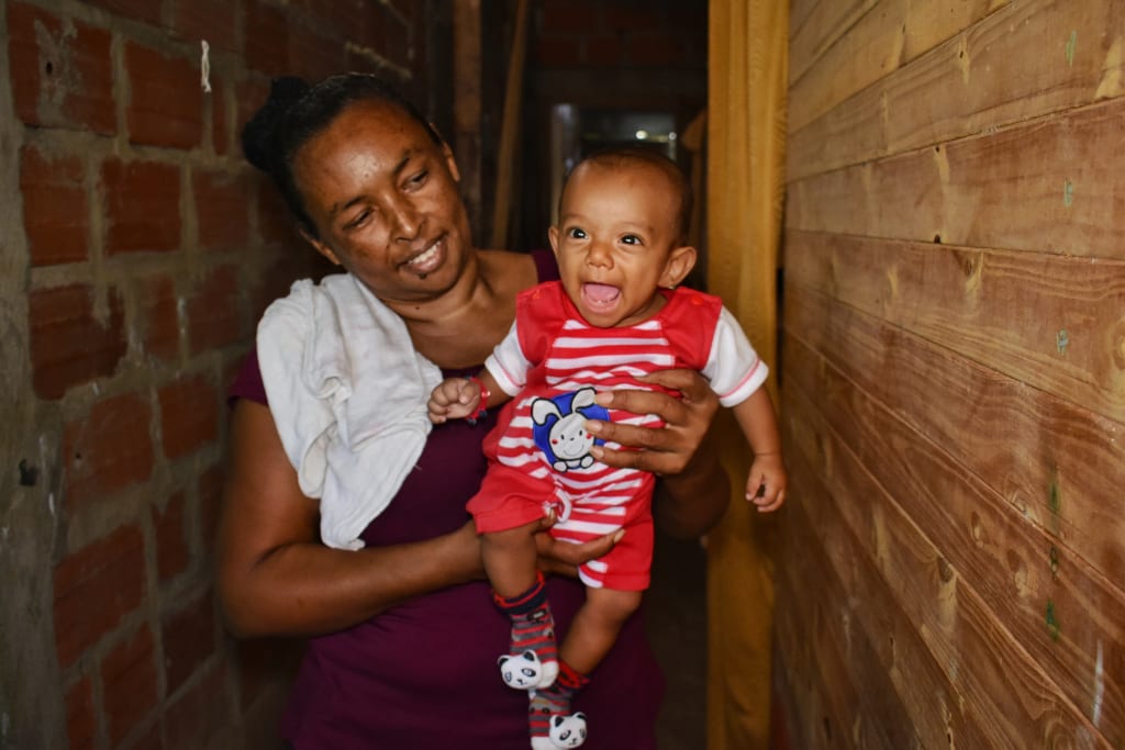 A woman with a white cloth over her shoulder holds a baby boy wearing red. They both smile.