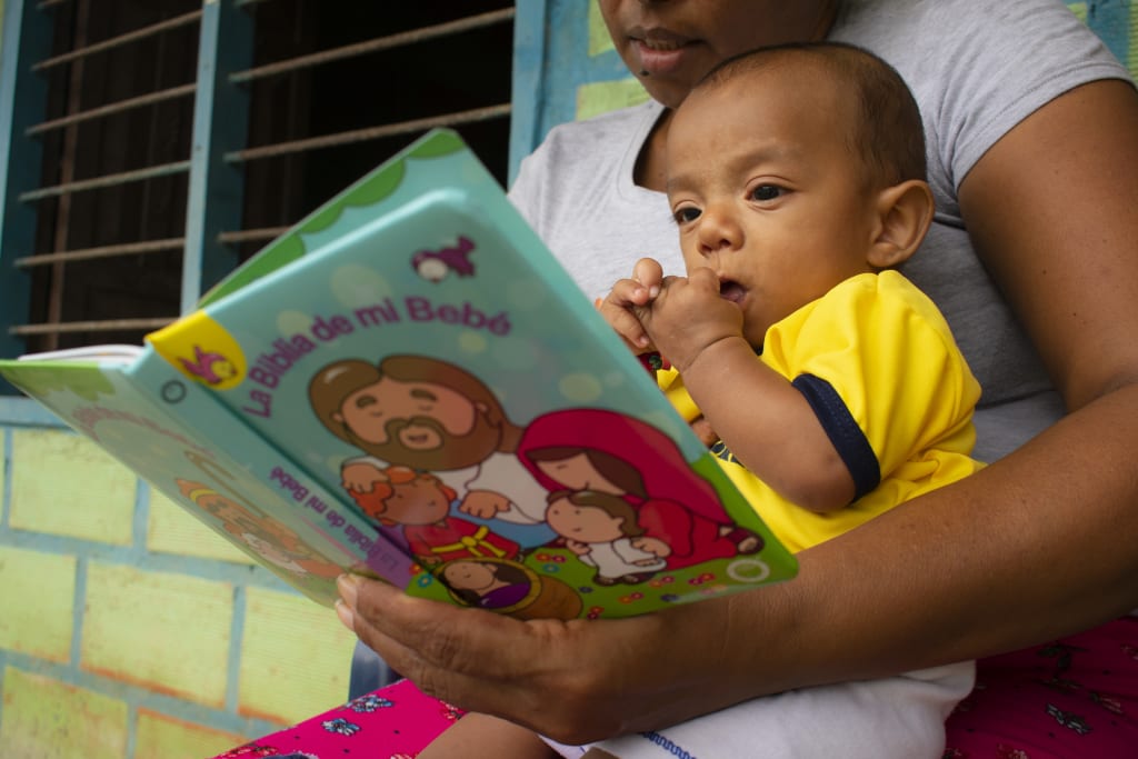 A baby boy in yellow sits on a woman's lap and looks at a picture book.