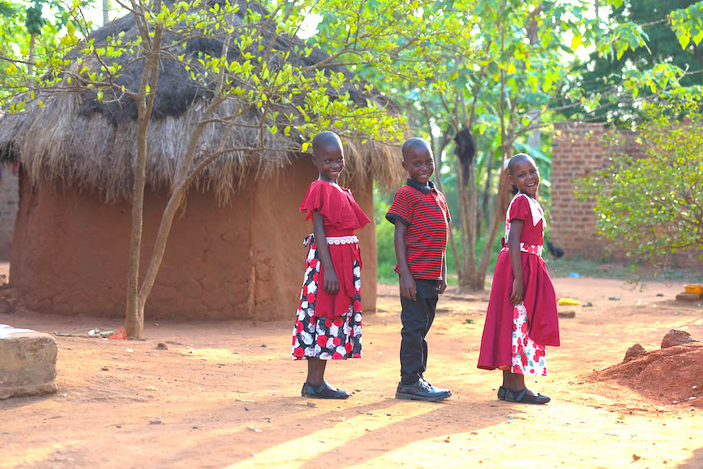 Triplets pose wearing red outfits by a hut. There are trees behind them and they are smiling. This pictures shows their development in 9 years!