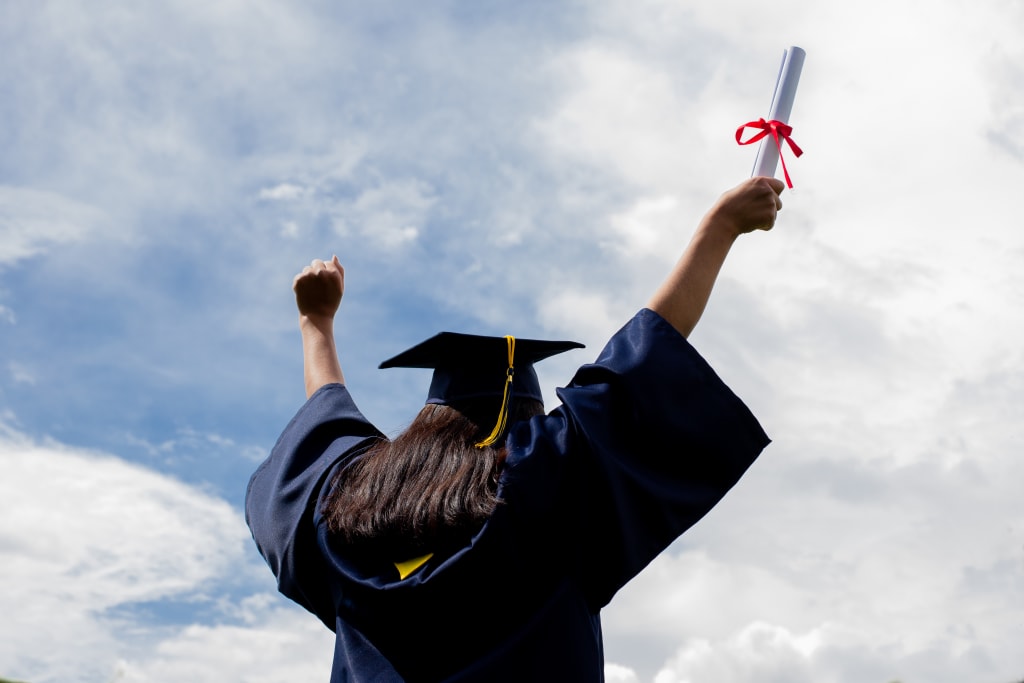 A young woman reaches her hands up to the sky, wearing a graduation cap and gown and holding a diploma