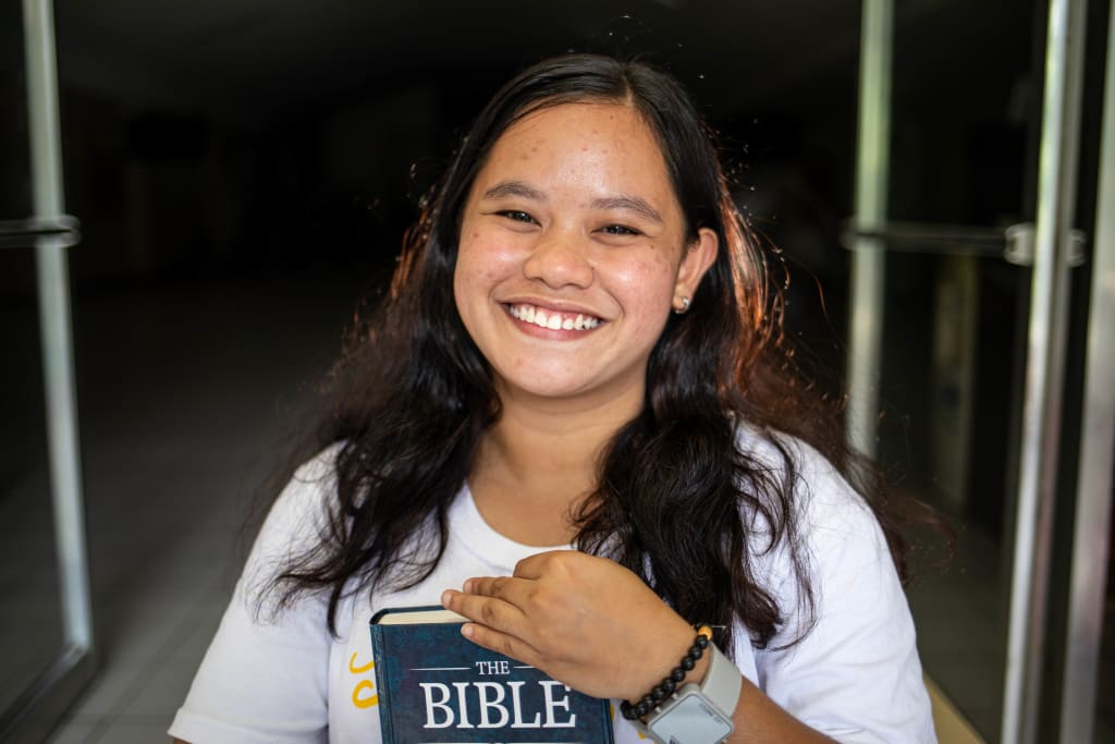 A young woman smiles brightly wearing a white shirt, with a Bible in her hand at the front steps of the church where she works. Jojin is preparing to become a teacher as she graduates from the Compassion program.
