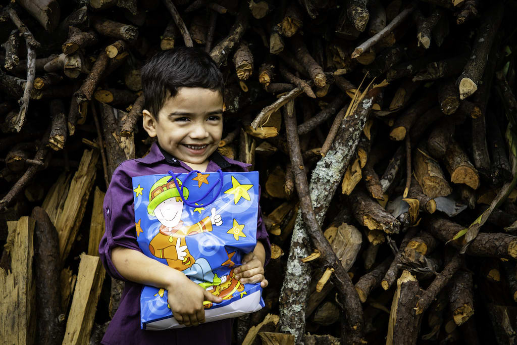 Estiven is wearing a purple shirt and dark pants. He is standing in front of a pile of firewood and is holding a gift he received from his sponsor.
