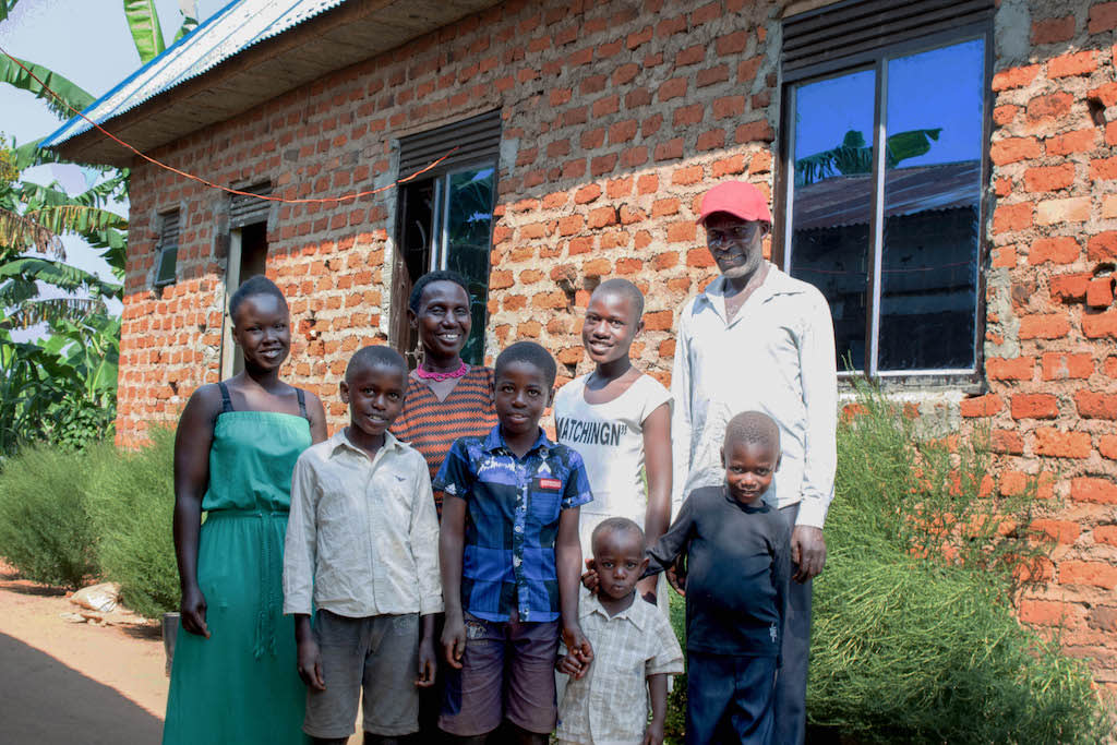 Racheal is standing with her family outside their new home, which is made of brick and has glass windows. Back row from left to right: Brenda (Racheal's aunt), Mary (Racheal’s grandmother), and Lauben (Racheal's grandfather). Front row from left to right: Racheal's cousins Hannan, Gilbert, Edison, and Alexander.