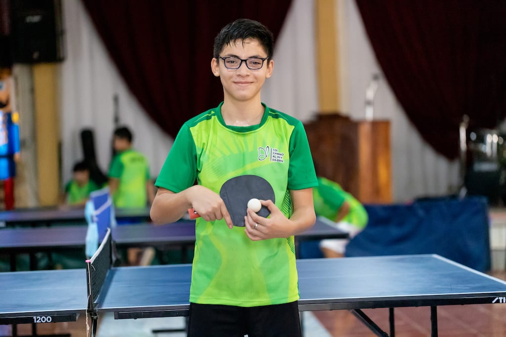 Young man smiles at the camera while holding a ping pong ball and paddle