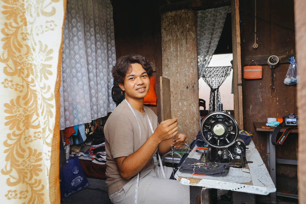 Julio is sitting at and using a sewing machine. He is in a small room in his house that he has turned into a sewing studio.